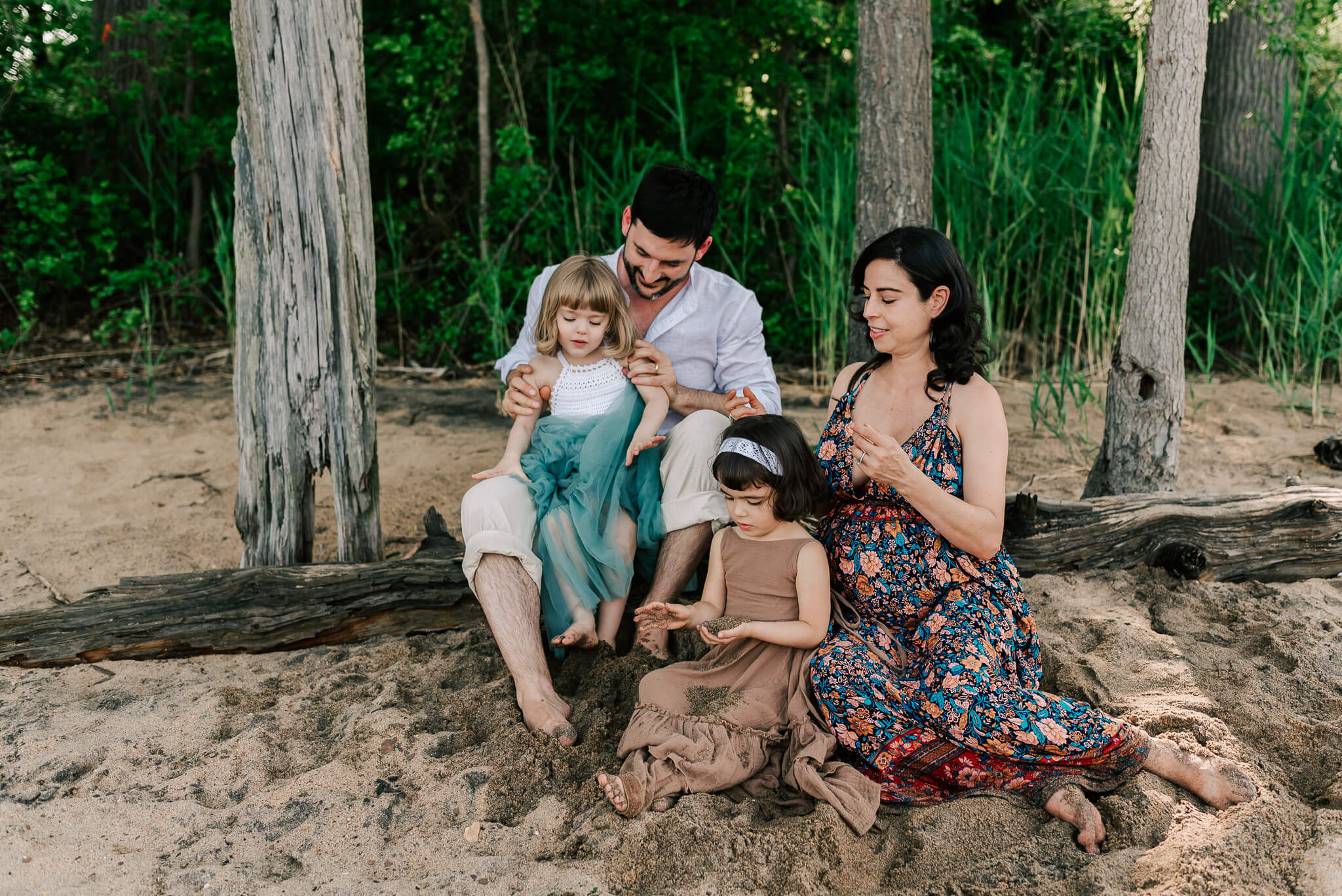 A pregnant mother plays with the hair of her daughter, sitting in the sand in front of her while dad sits on a log with their other daughter in his lap