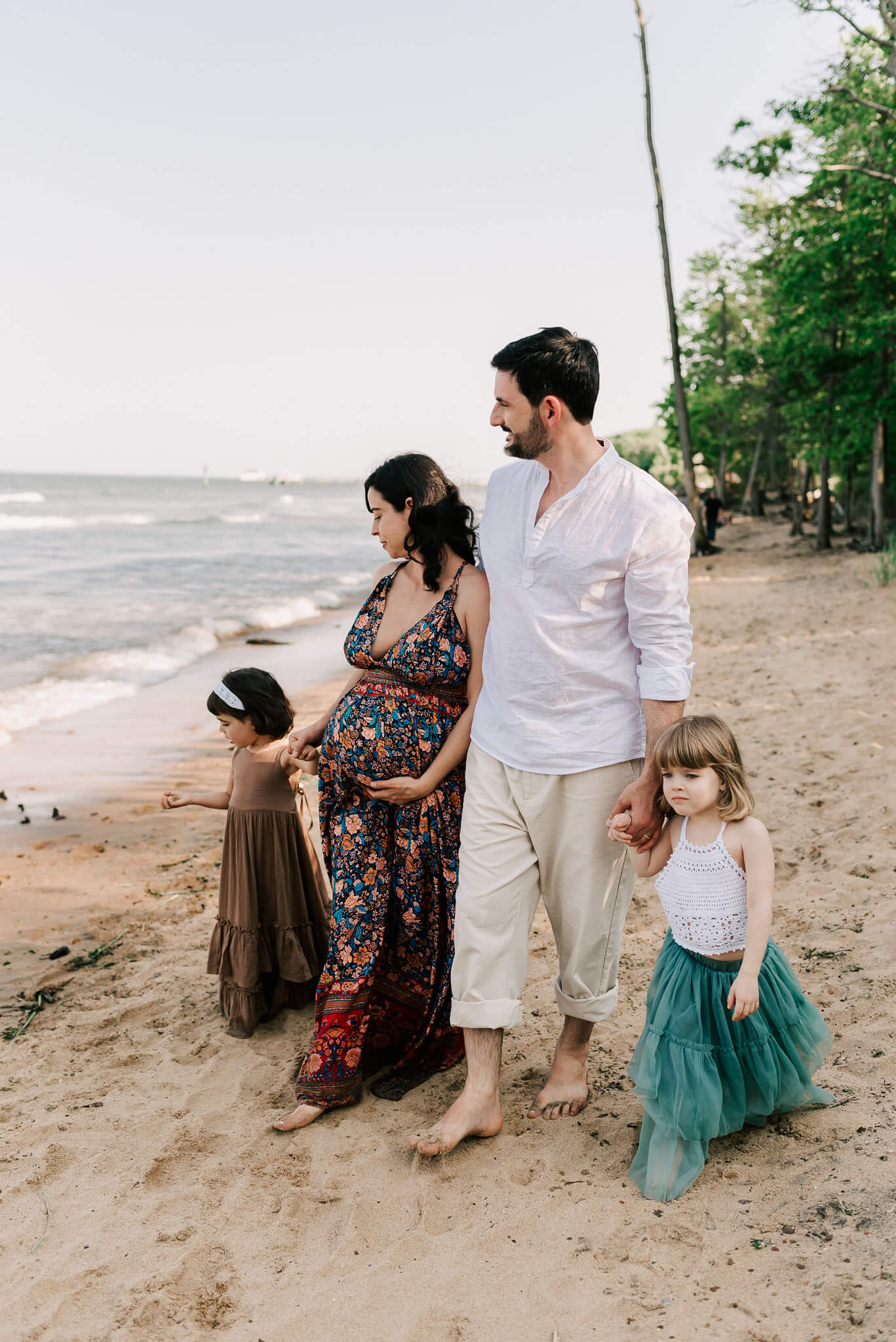 A family of four walk down a sandy beach lined with trees while mom holds her pregnancy bump kid to kid centreville