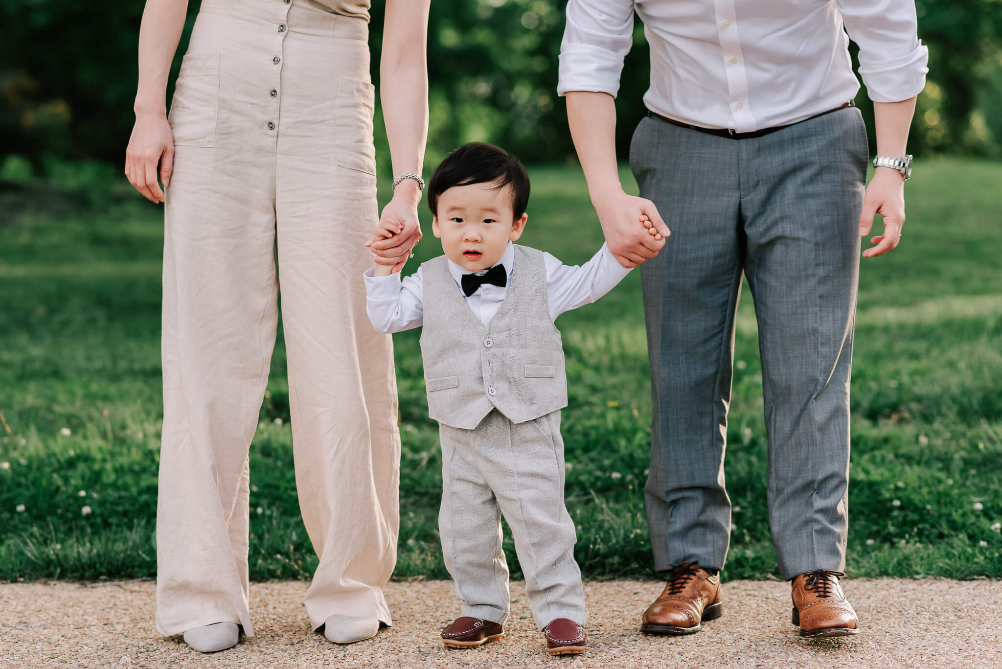 A toddler boy stands holding parents hands in a park path wearing a grey suit and black bowtie