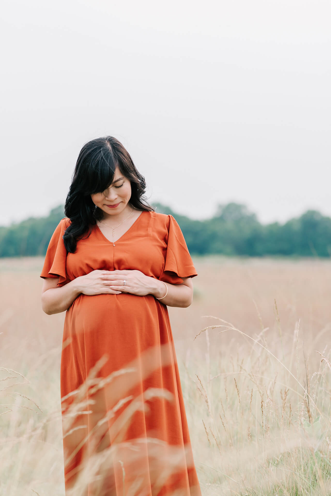 A mom to be stands in a field of tall grass looking down at her bump in a red maternity gown