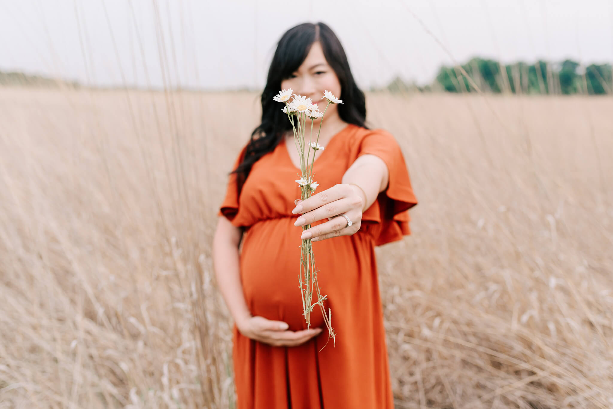 A mother to be stands in a field of tall golden grass in a red maternity dress holding a handful of wildflowers in front of her alexandria midwives