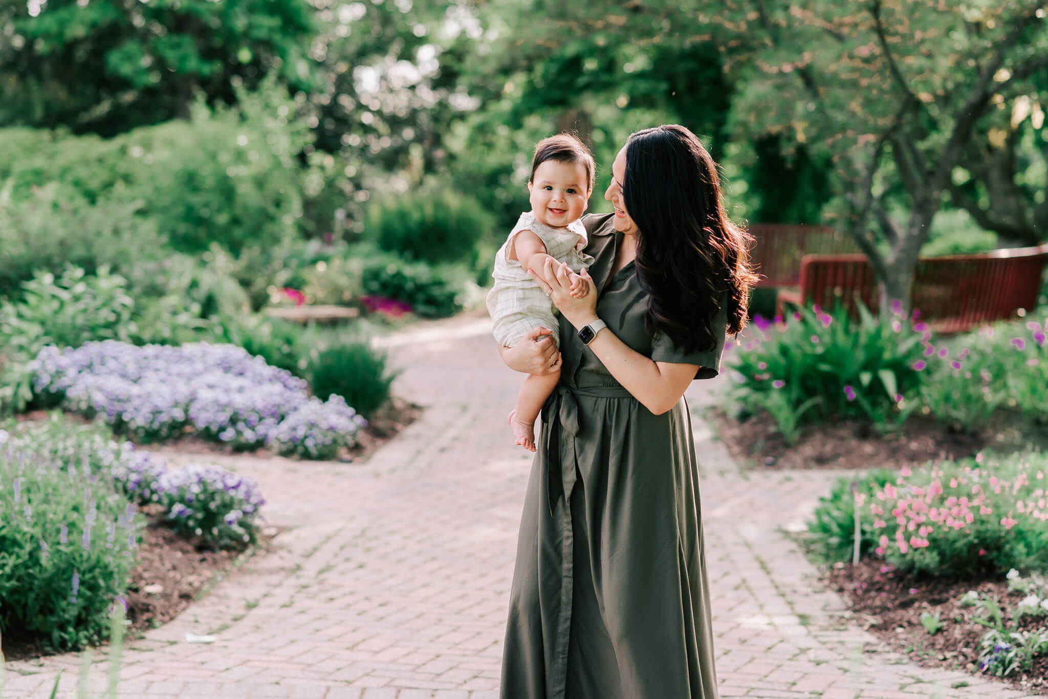 A mother in a dark green dress stands in flower garden path playing with her infant child on her hip Capital Women’s Care Fairfax