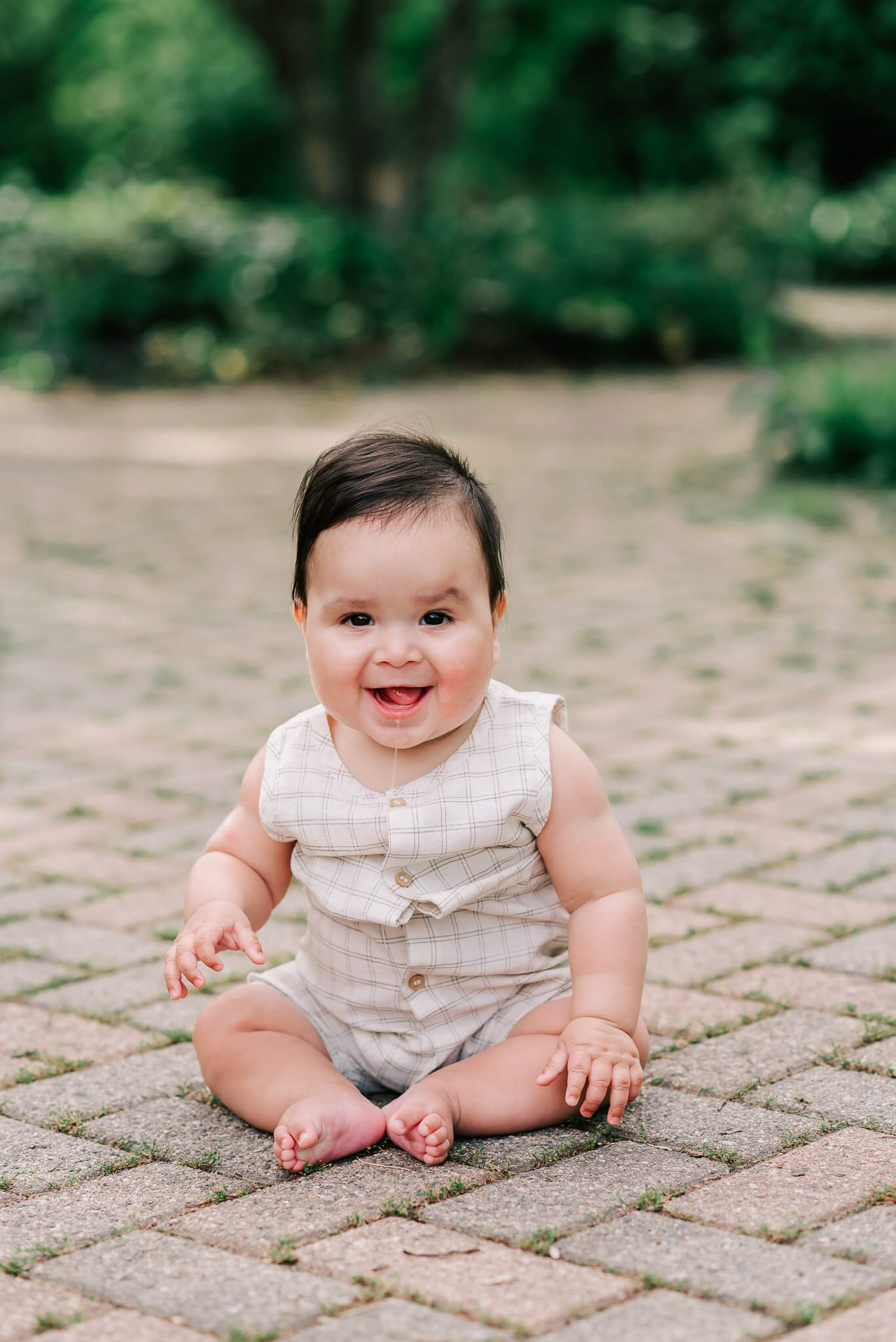 An infant sits in a plaid onesie drooling and smiling on a brick path in a park Capital Women’s Care Fairfax
