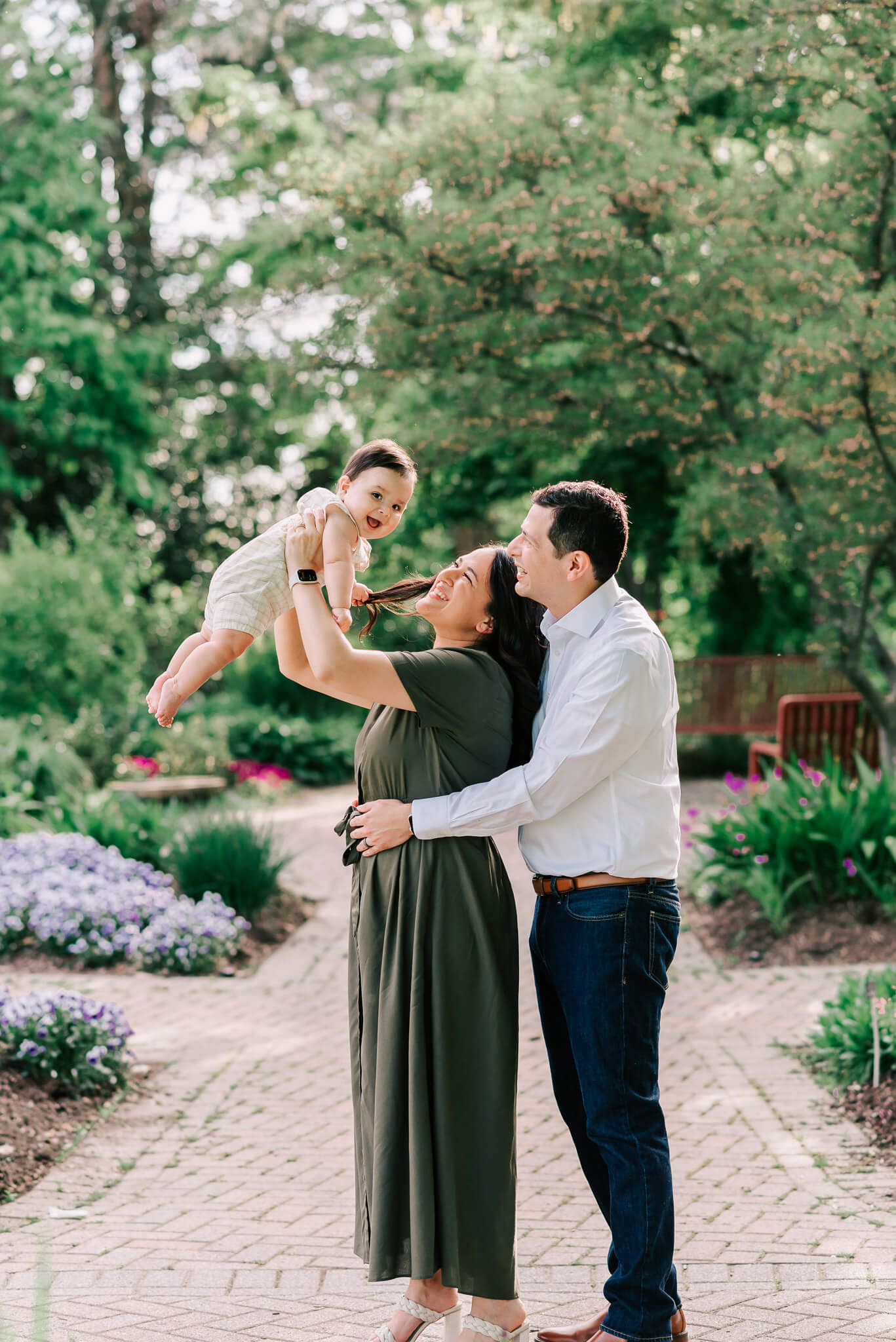 A mother and father lift and play with their infant child while standing in a flower garden path