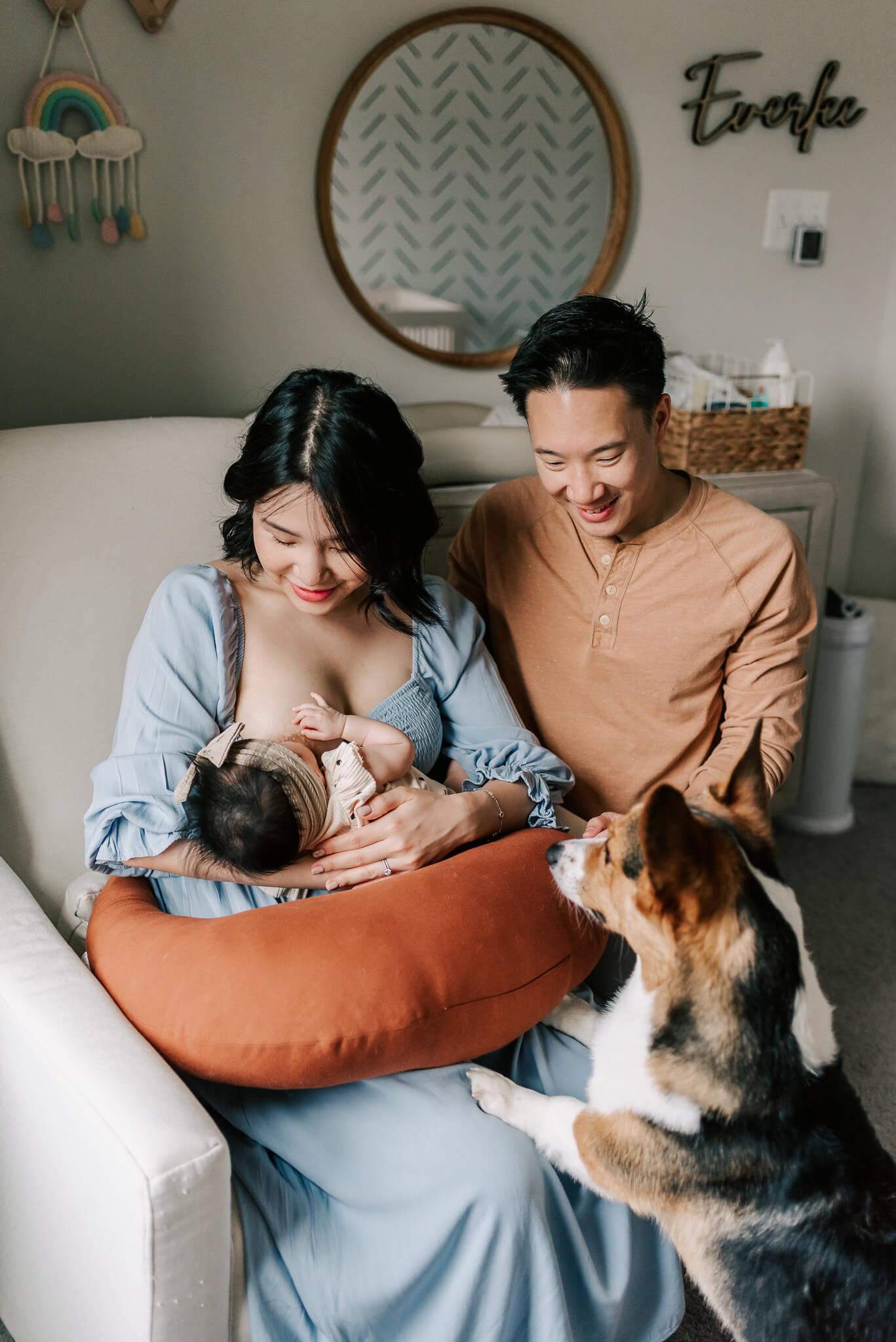 A mother breastfeeds her newborn baby while sitting in a nursing chair in a blue dress while dad and their corgy puppy lean in