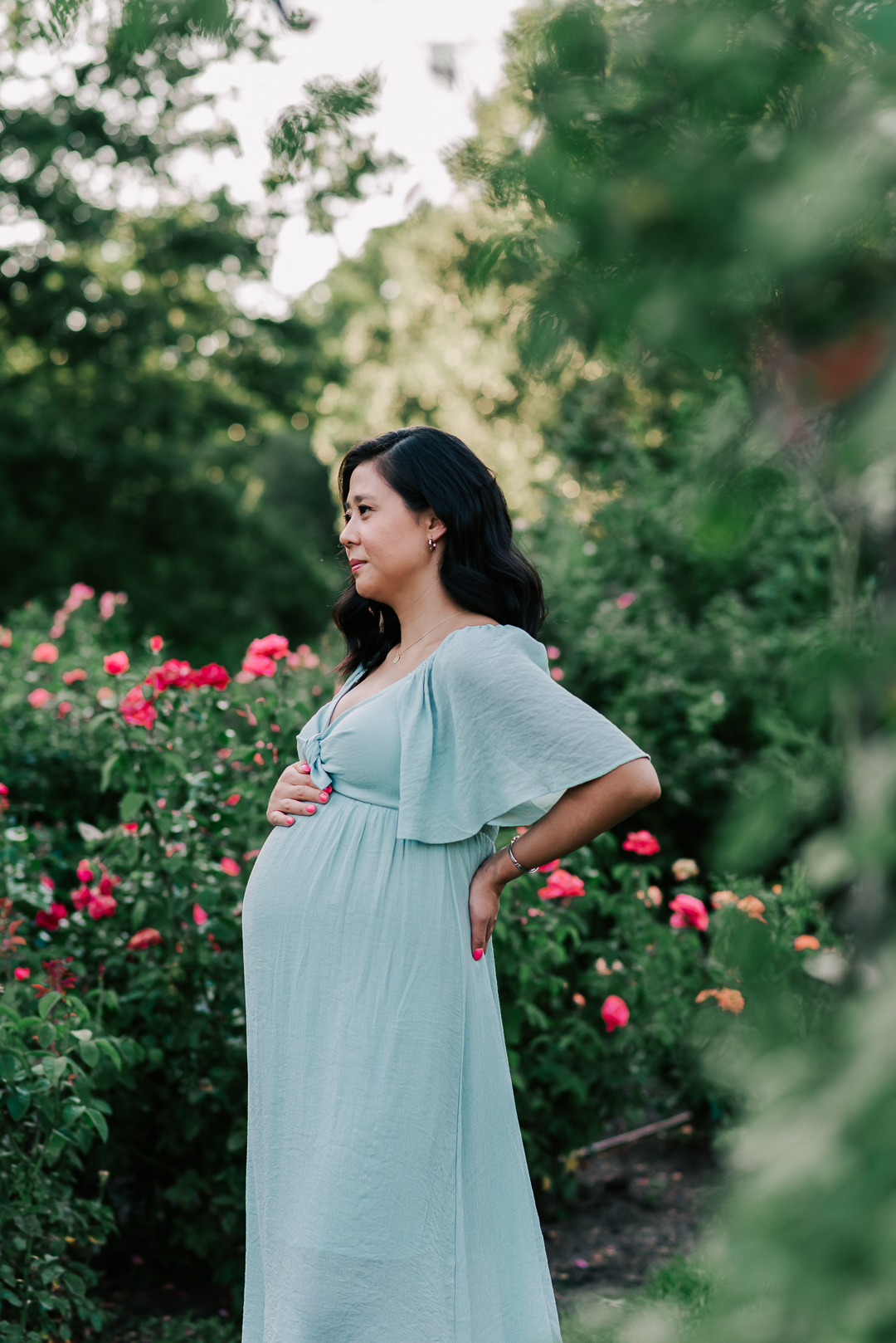 A mom to be stands with a hand on her bump and hip while standing and smiling in a rose garden