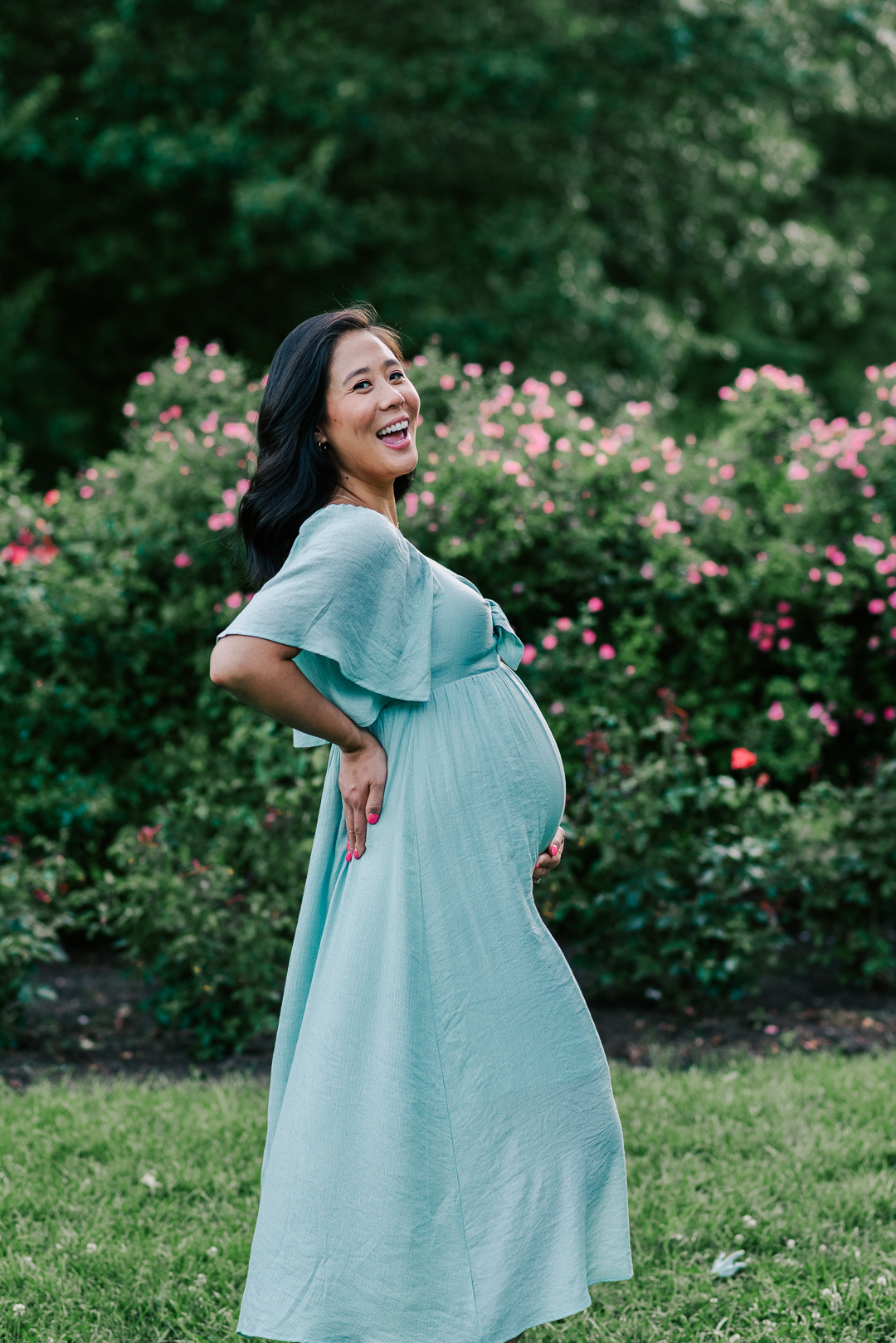 A mom to be laughs while holding her bump and standing in a rose garden doulas of capitol hill