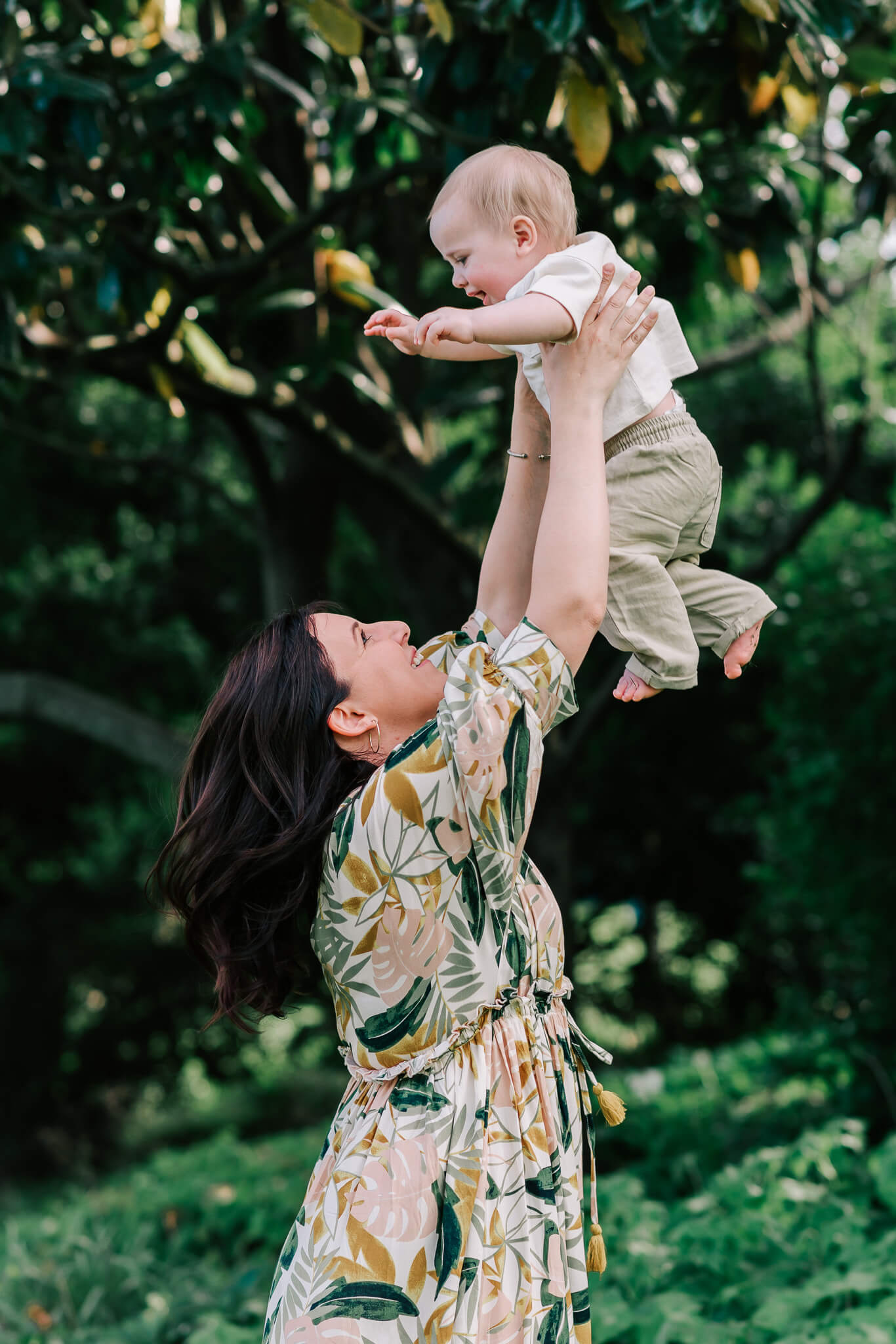 A mother in a tropical print dress lifts her young child in the air to play in a garden happy parents happy babies