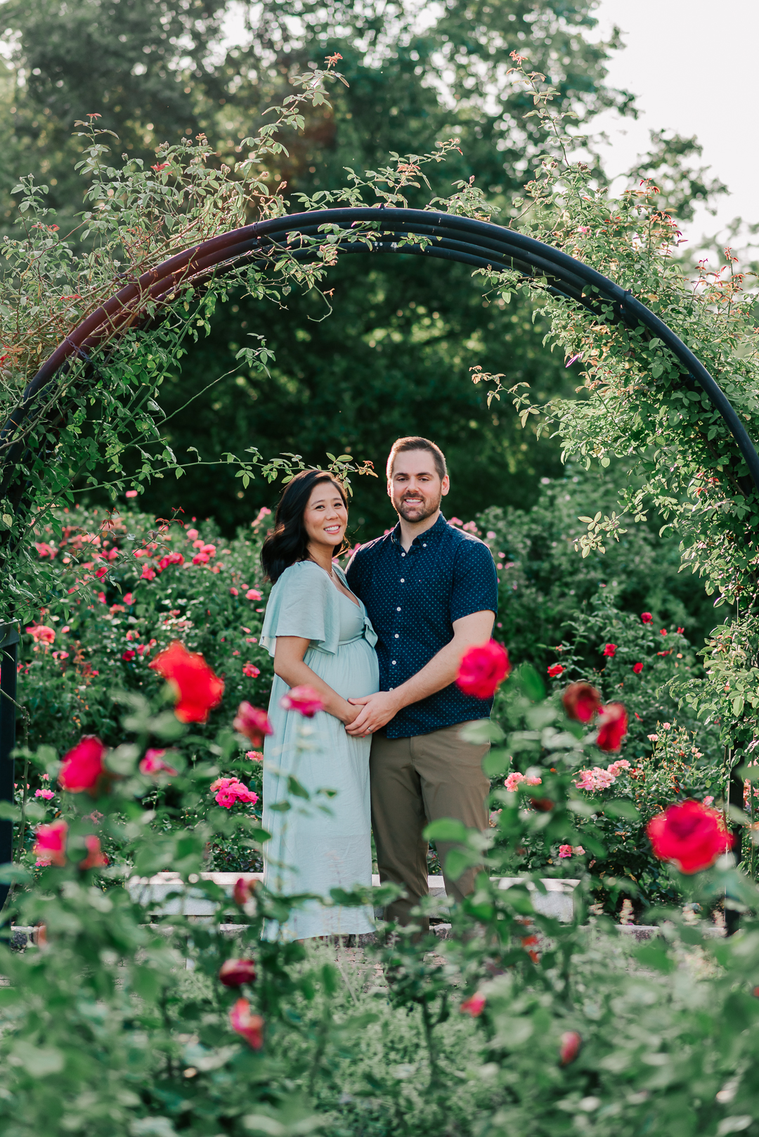 A mom to be in a blue dress stands in a rose garden under a large arch while hugging onto her partner the physician midwife collaborative practice