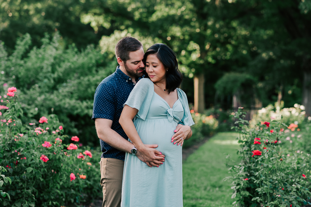 A mom to be leans into her partner with hands on the bump while standing in a rose garden the physician midwife collaborative practice