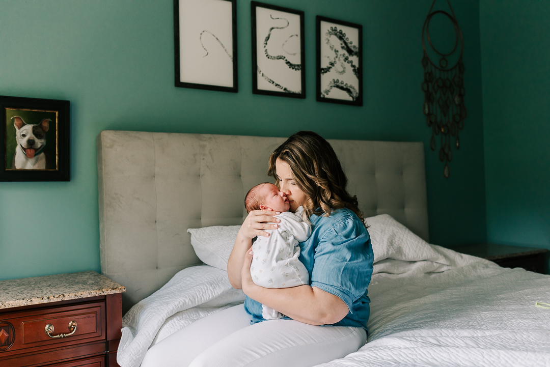 A new mom sits on a bed snuggling with her newborn baby in a blue top and white pants washington dc baby stores