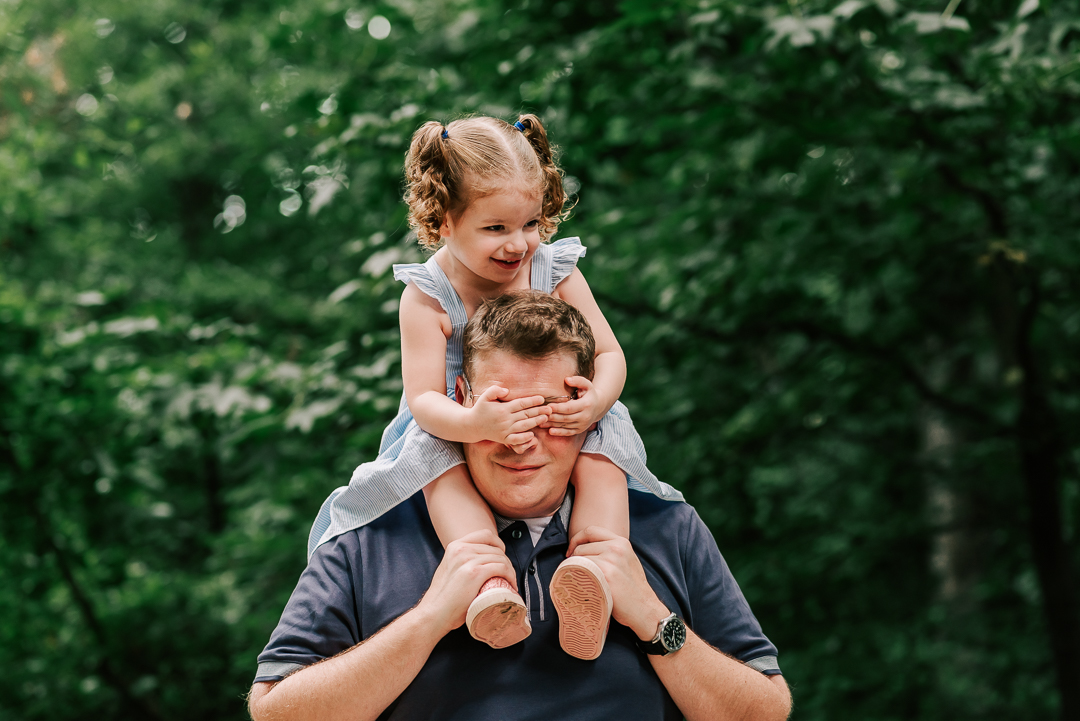 A toddler girl in a blue dress sits on dad's shoulders in a park and covers his eyes as she laughs