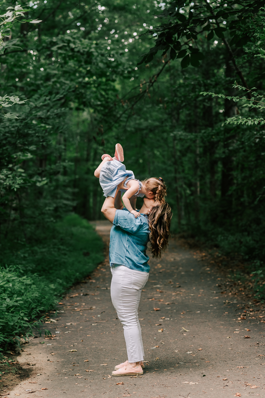 A mother stands in a wooded park path while lifting and kissing her toddler daughter in a blue dress above her head after visiting a centreville chriopractic center