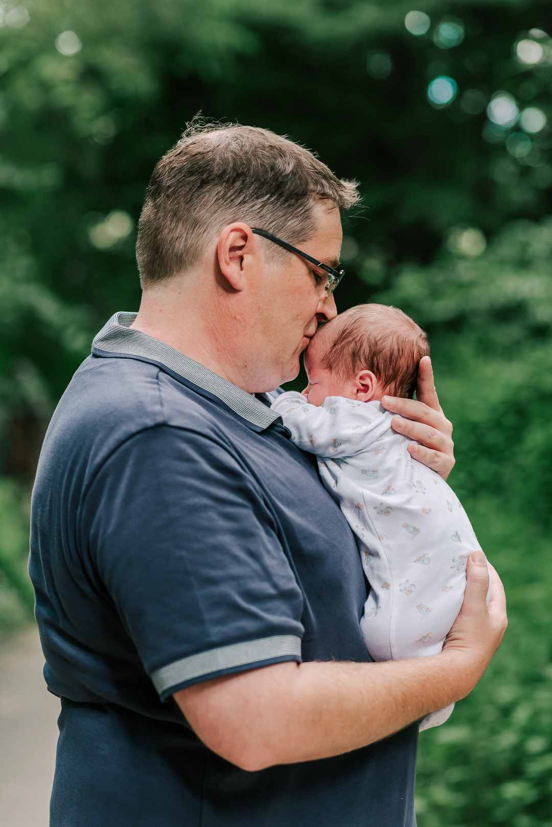 A father cradles his newborn baby on hist chest while kissing its head and standing in a park after visiting a centreville chriopractic center