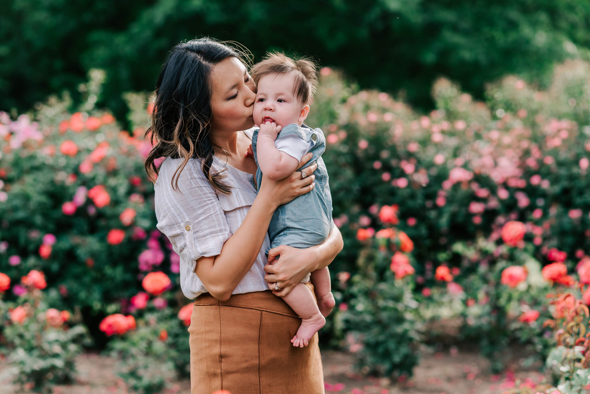 A mother in a tan skirt and grey top kisses the cheek of her infant baby in blue overalls while standing in a rose garden centreville obgyn