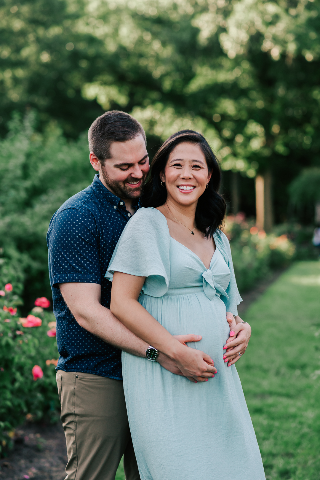 A mom to be leans into her husband as they both hold the bump while standing in a rose garden