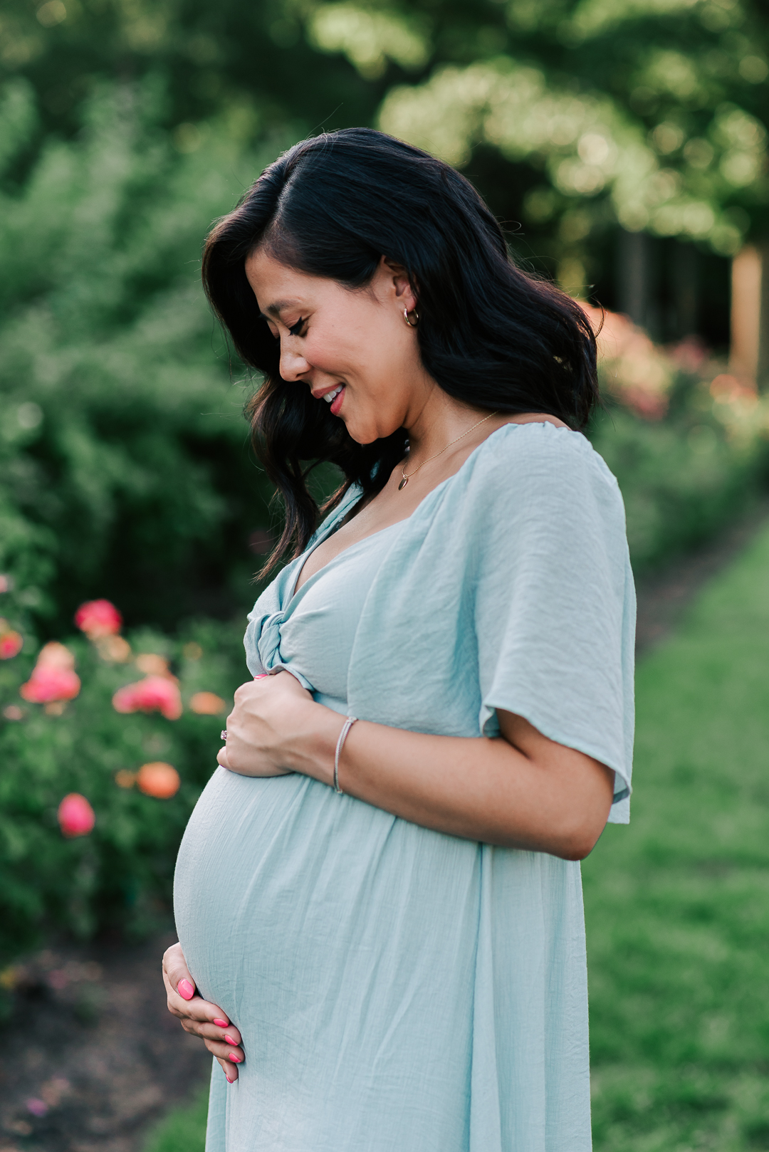 A mom to be stands in a rose garden smiling down to her bump in a blue maternity gown spas in northern virginia
