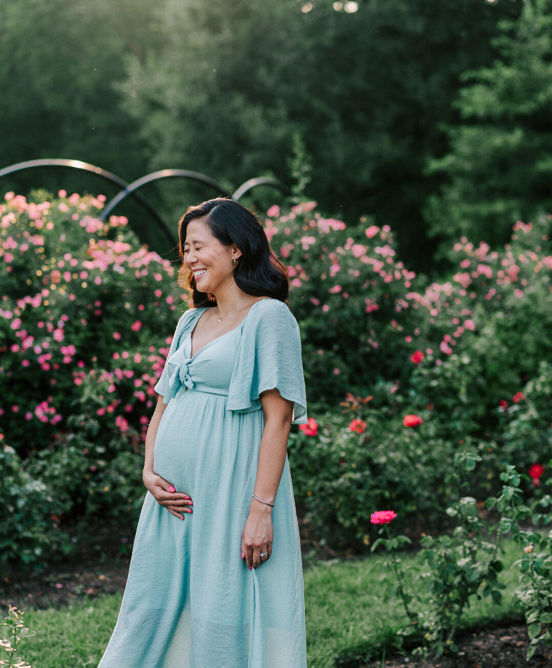 A mom to be laughs and holds her bump while standing in a rose garden spas in northern virginia