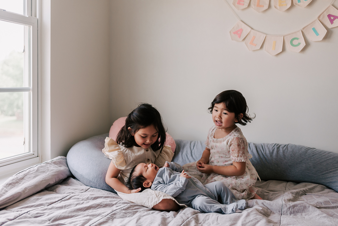 Two toddler girls in dresses sit on a bed by a window playing with their newborn baby brother in a blue onesie in one of their laps 529 kids consign