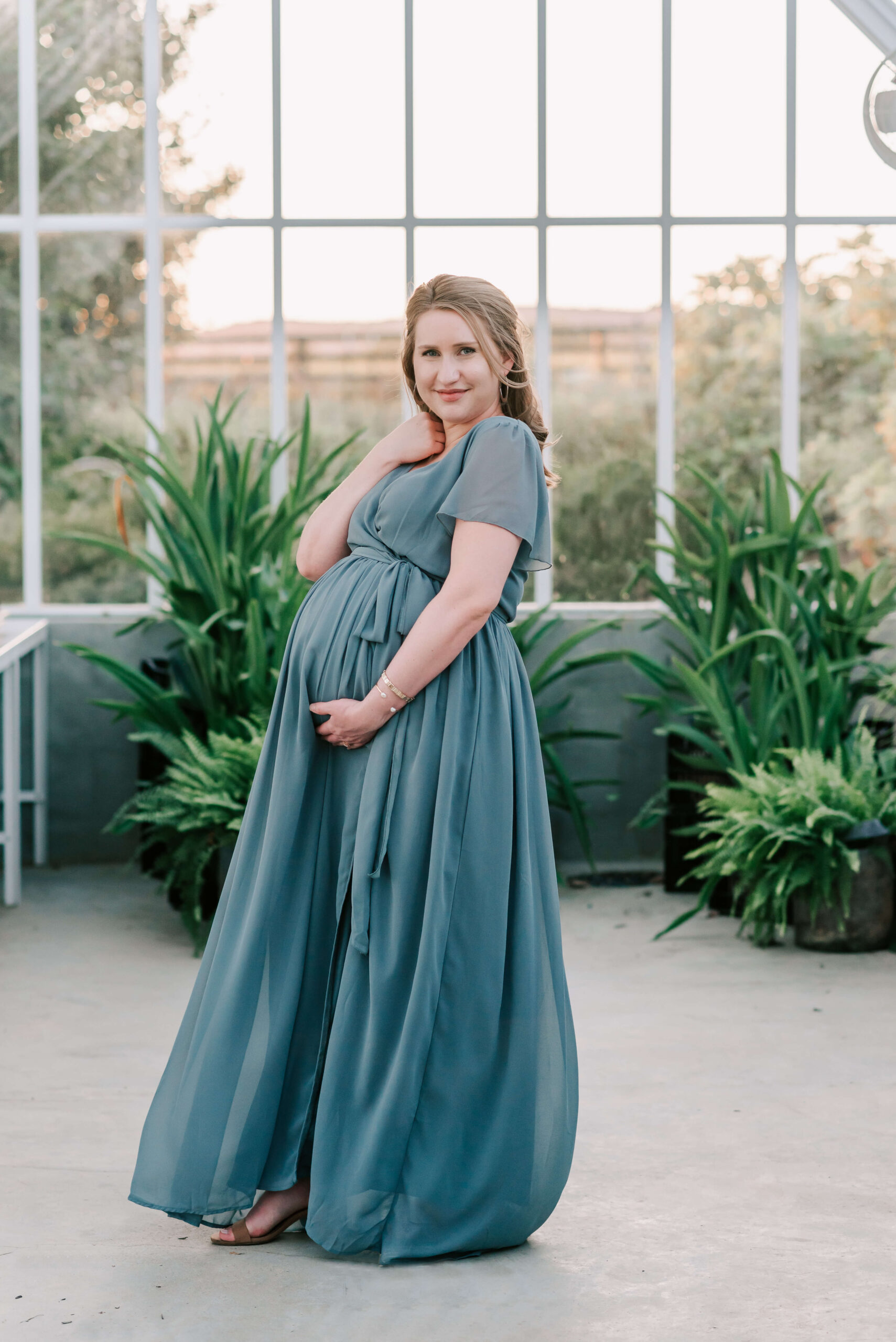 A mom to be holds her bump while standing in a green house wearing a blue maternity gown