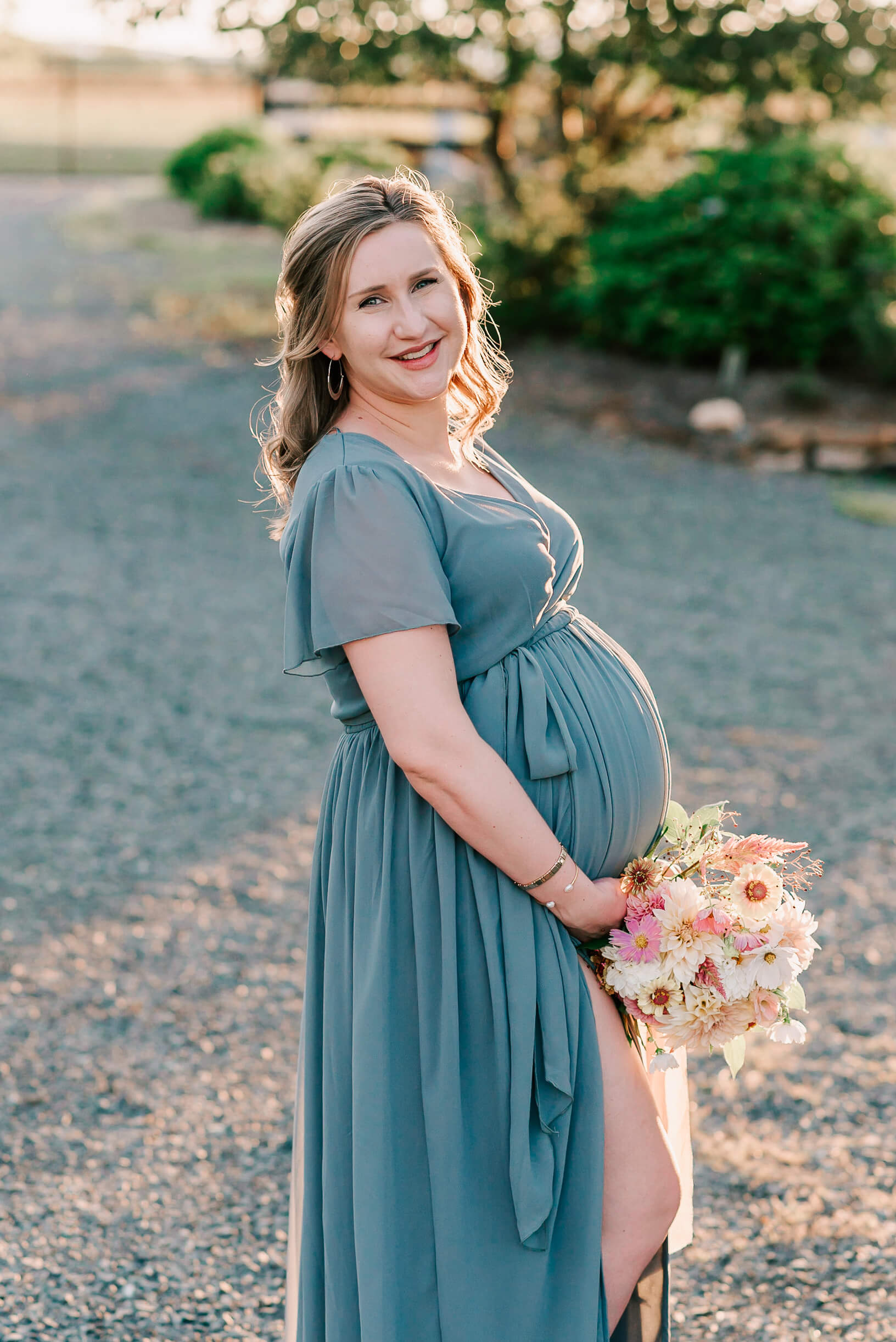 A mom to be holds a white and pink bouquet while holding her bump and standing in a garden path at sunset before getting gifts from bellies & babies
