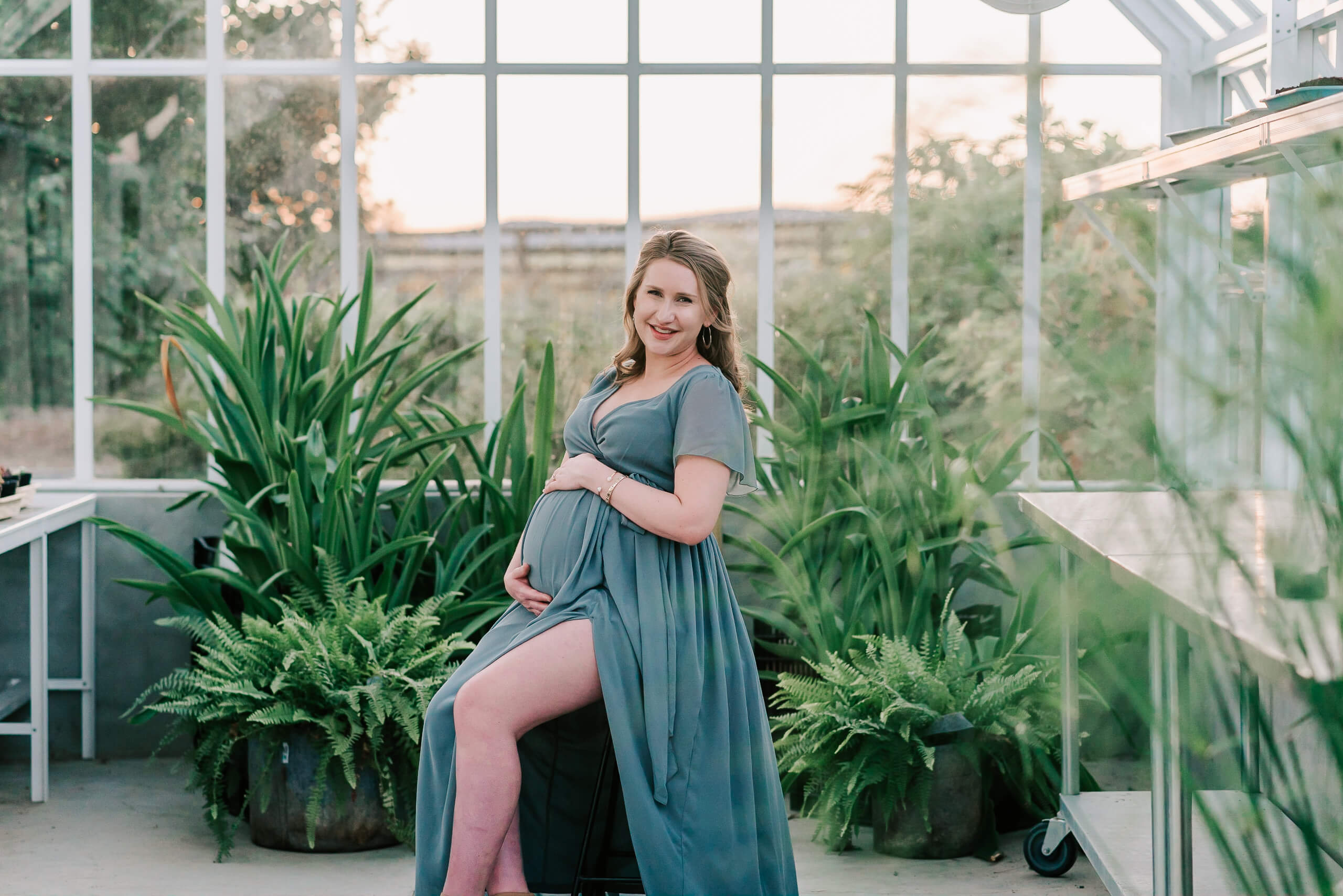 A mom to be in a blue maternity gown sits on a stool in a greenhouse holding her bump bellies & babies