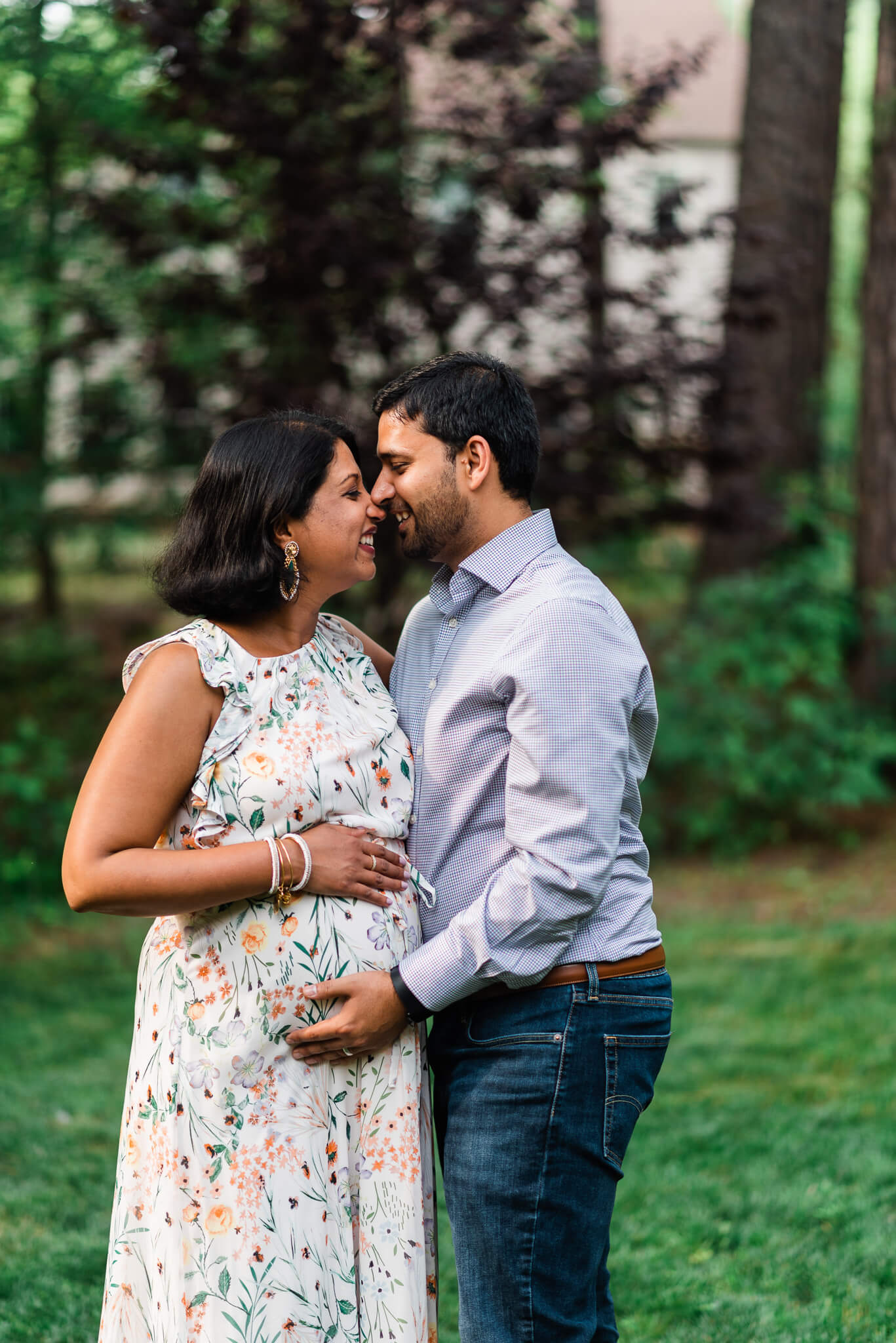 A mom to be leans in for a kiss from her husband in a floral dress while standing in a park holding the bump