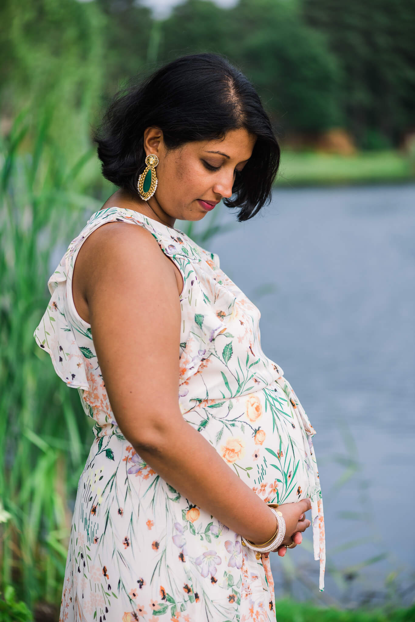 A mom to be gazes down at her bump taken care of by center for midwifery while standing on the edge of a lake