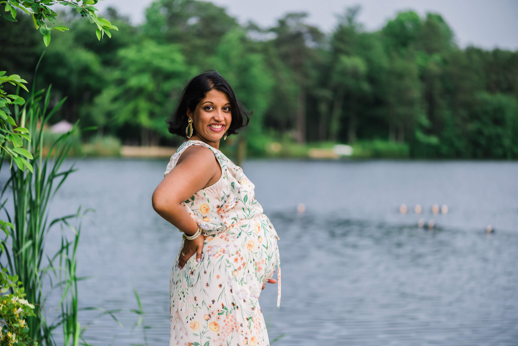 A mom to be in a white floral dress stands by a lake holding her bump in a park after meeting with center for midwifery