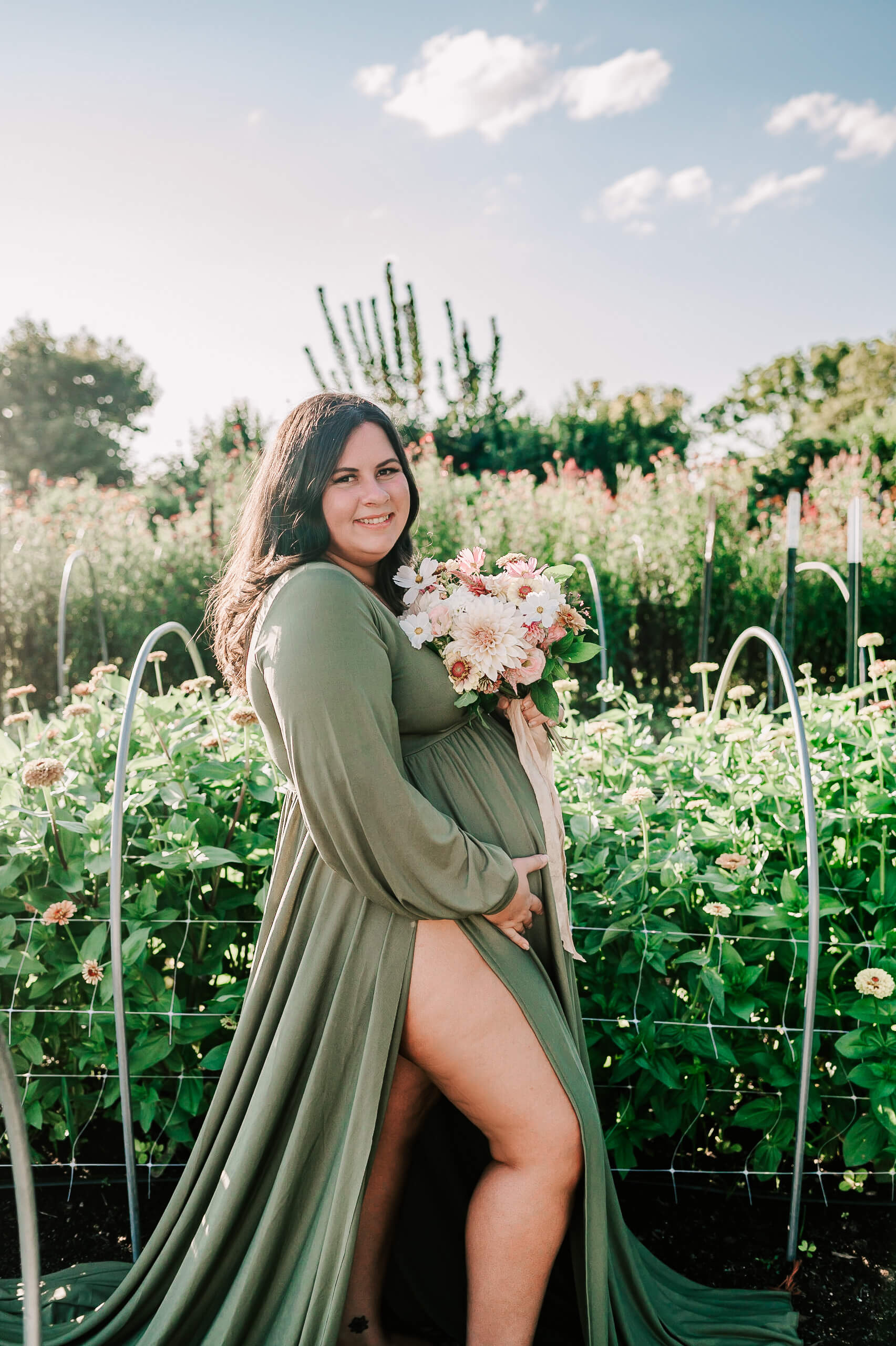 A mom to be in a green maternity gown holds a bouquet of pink and white flowers while standing in a flower garden