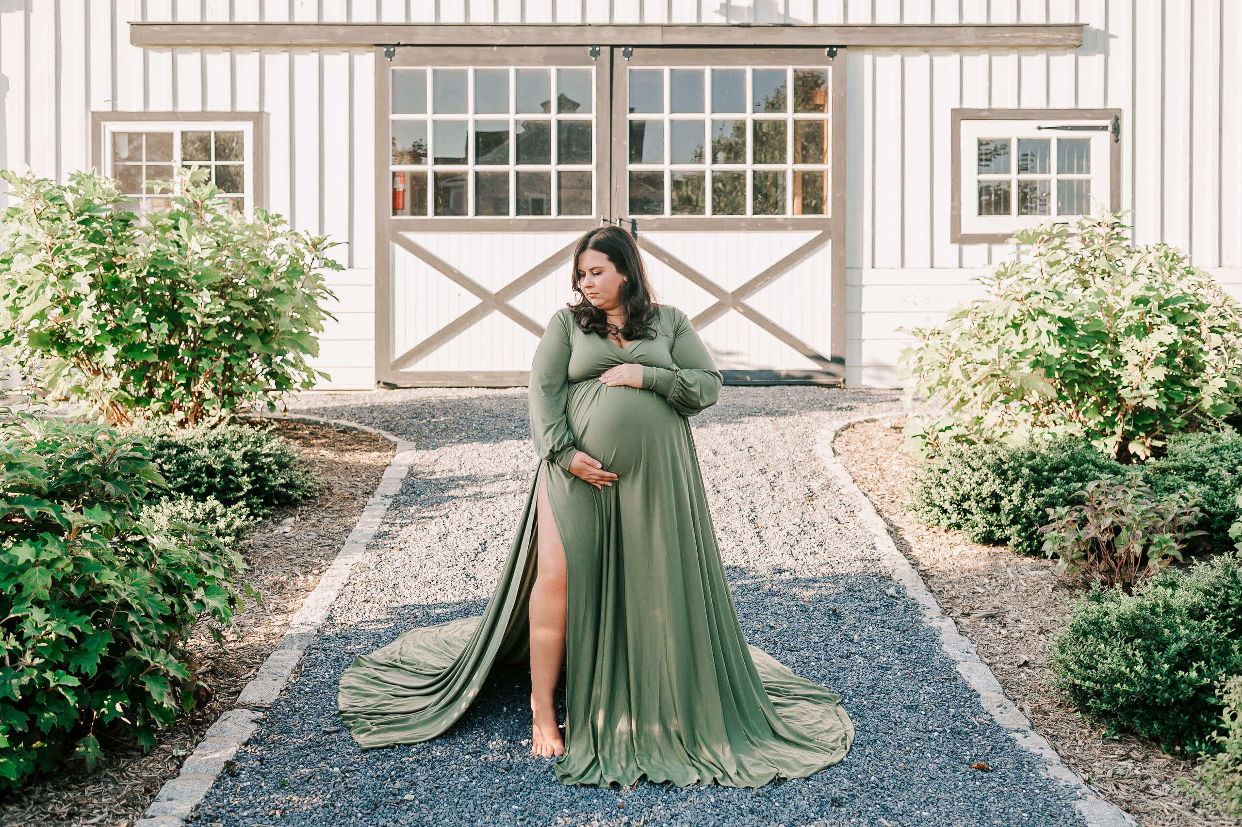 A mom to be holds her bump in a green maternity dress while standing in a garden path outside a white barn before visiting community of hope birth center