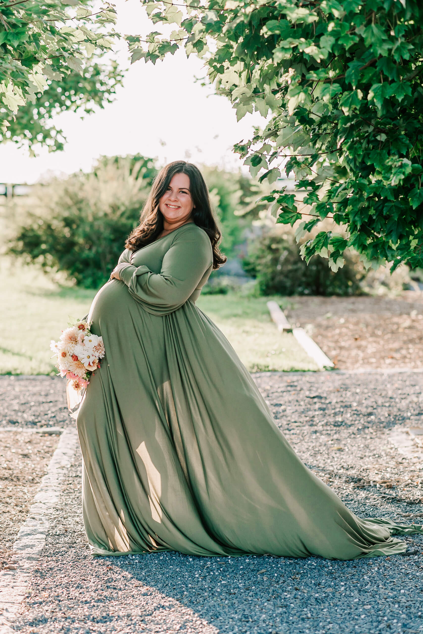 A mom to be stands in a park wearing a green maternity gown blowing in the wind and holding a bouquet of flowers after visiting community of hope birth center