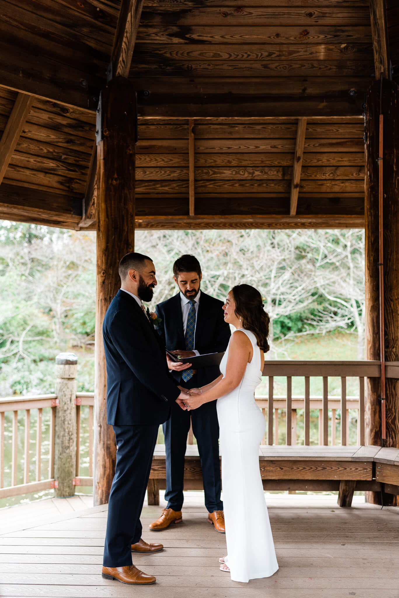 Newlyweds stand under a gazebo holding hands during their wedding ceremony smiling at each other