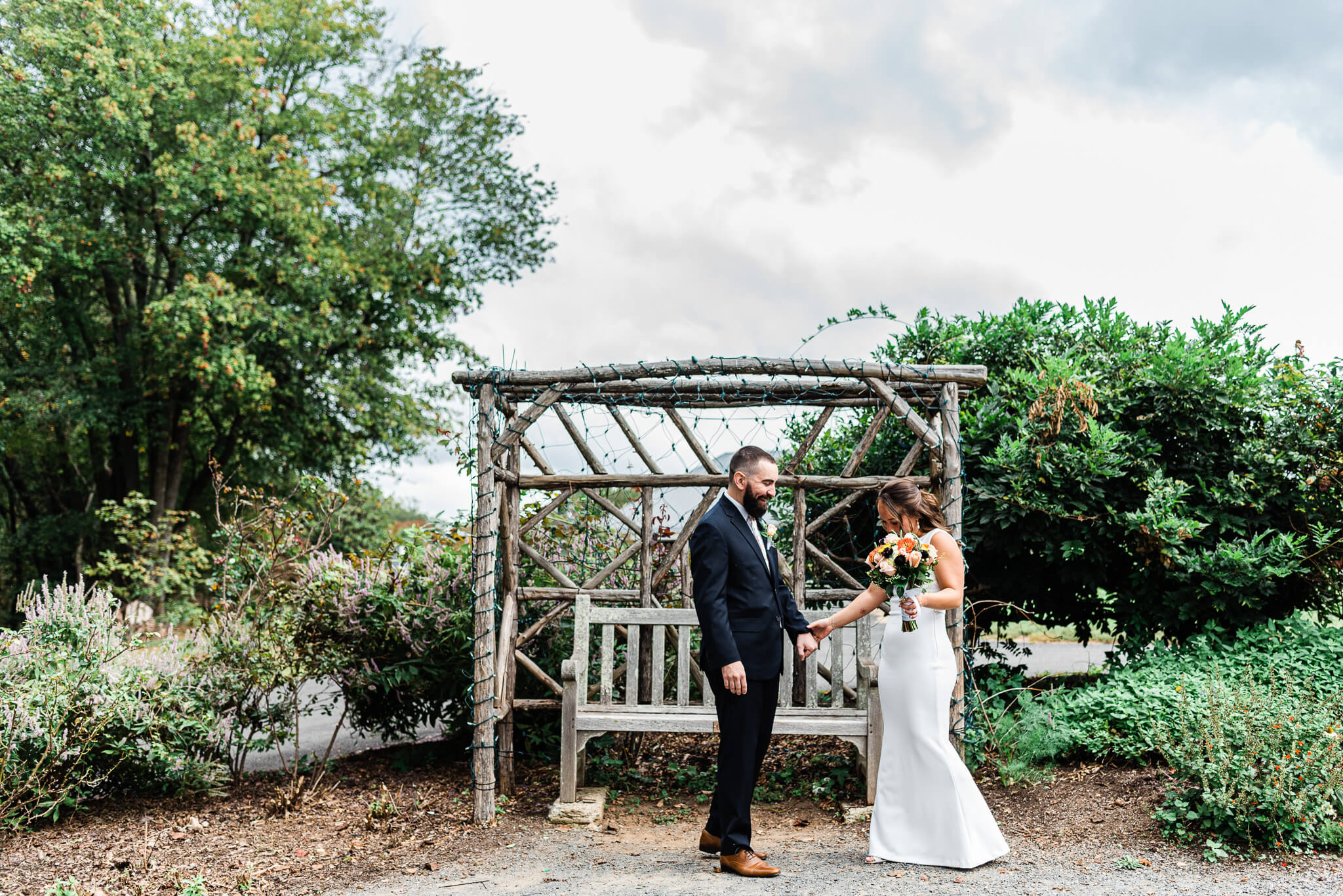 A groom in a black suit helps his bride walk through a garden holding her colorful bouquet by a wooden bench and pergola