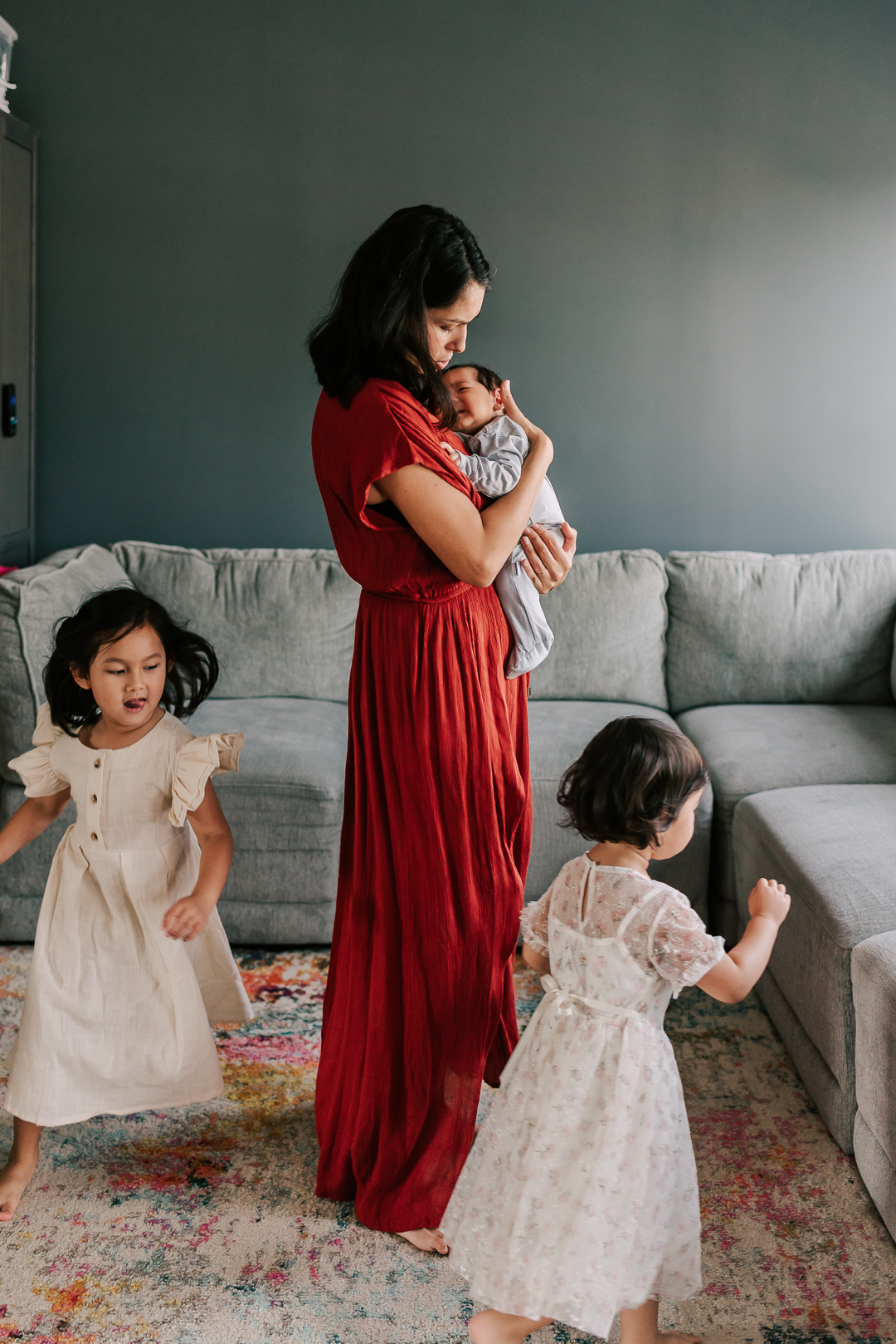 A mom in a red dress stands in a living room cradling her newborn baby while her two young daughters play around her