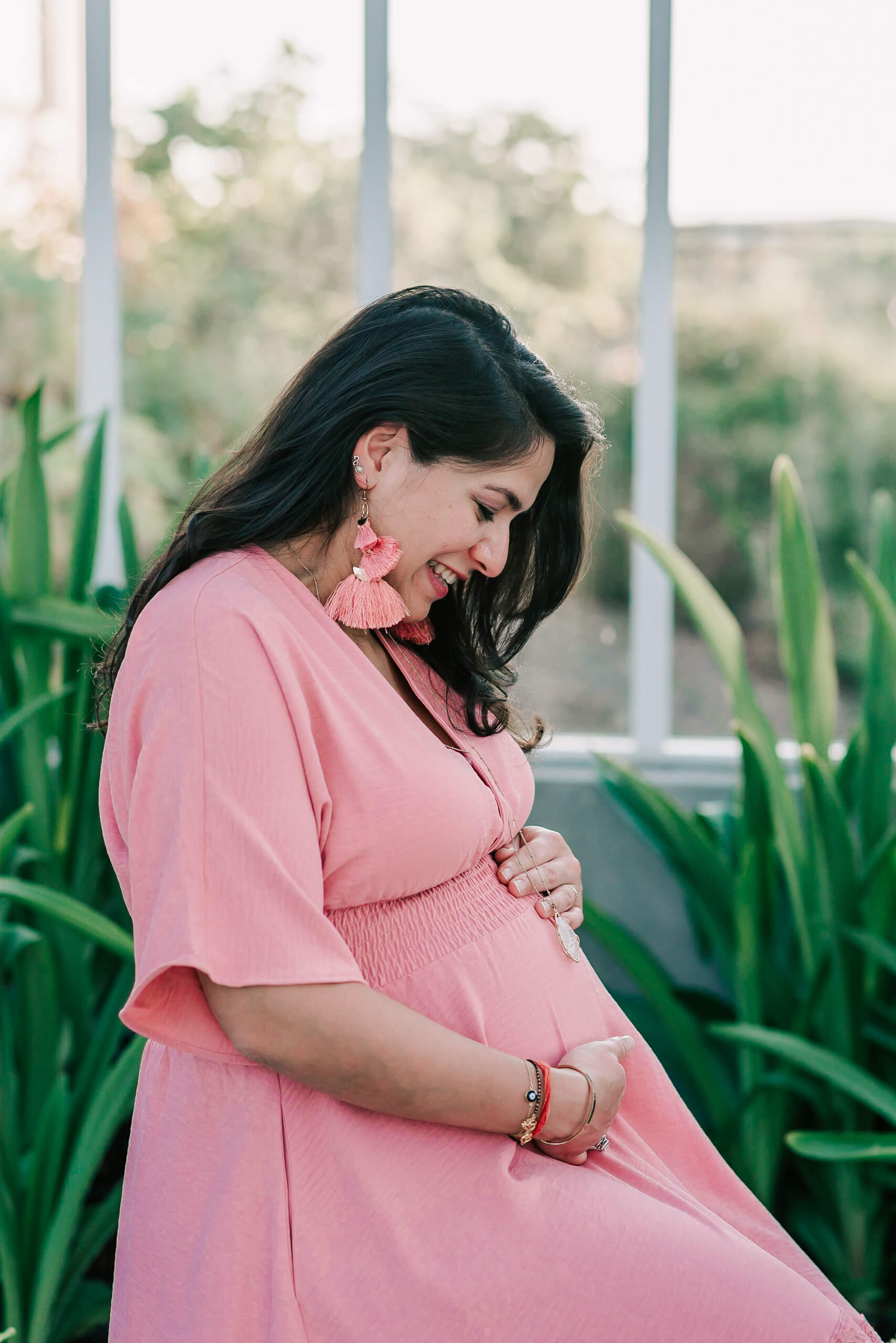 A mom to be sits on a stool in a greenhouse while holding and smiling down to her bump