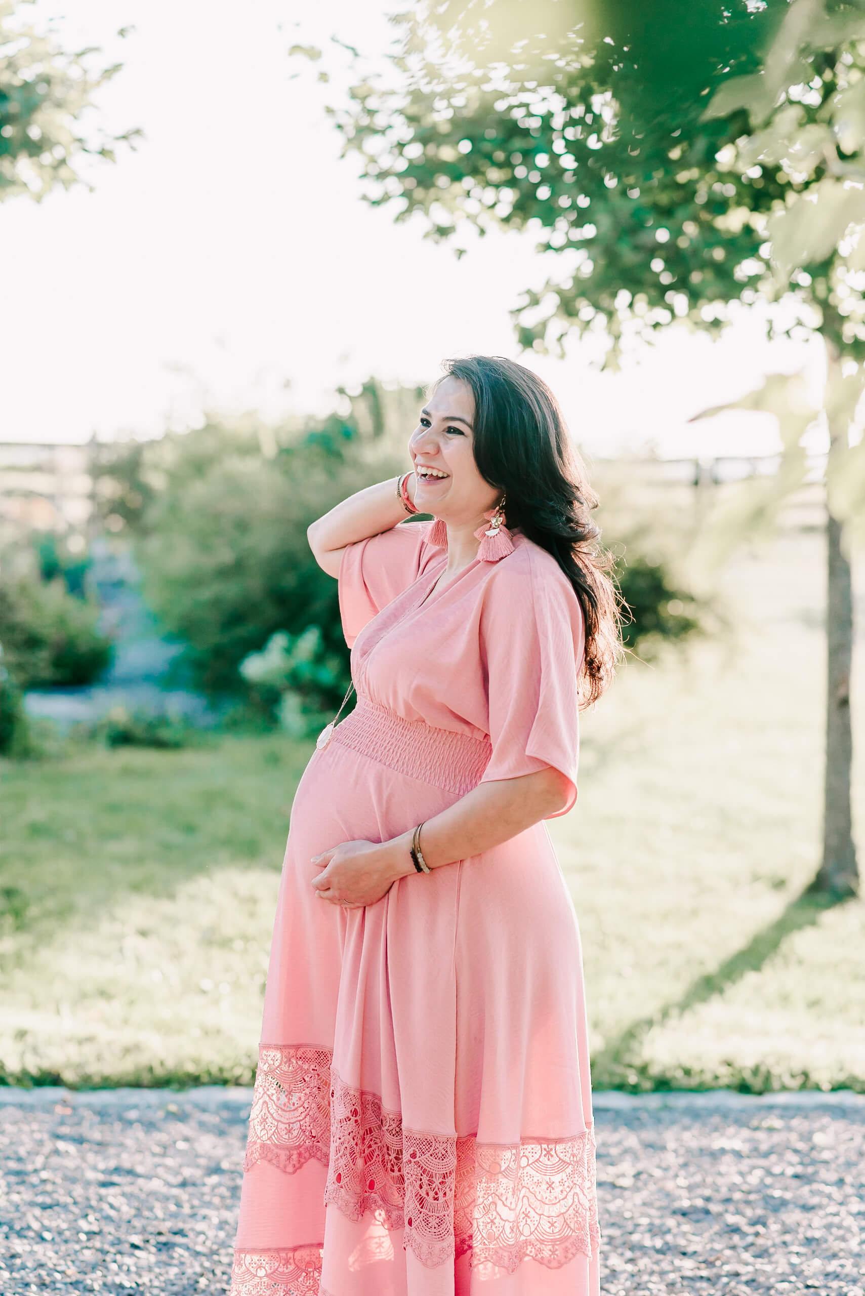 A mother to be stands in a park path in a pink maternity dress with a hand pulling some hair back and smiling before visiting virginia hospital center arlington