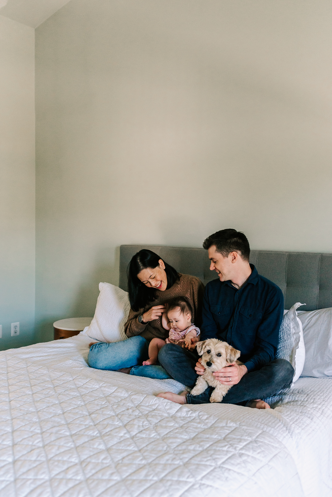 A mom and dad play with their infant daughter and small dog on a bed after having a fairfax home birth