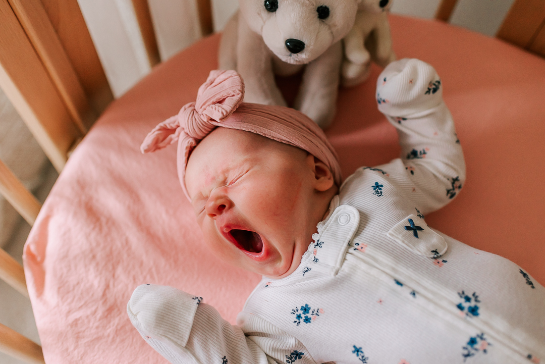 A newborn baby yawns while laying in a crib in a floral onesie after visiting inova obgyn falls church