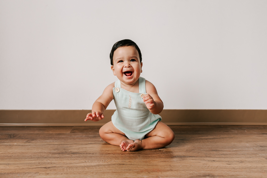 A happy toddler in green overalls sits on the floor of a studio