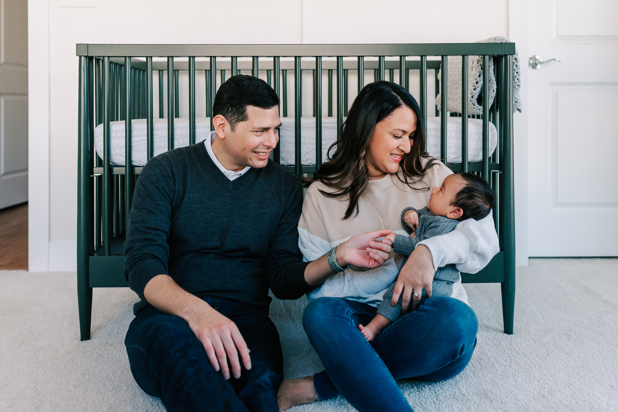 A mom and dad sit on the floor against a green crib playing with their newborn baby in mom's lap thanks to OrthoPelvic PT