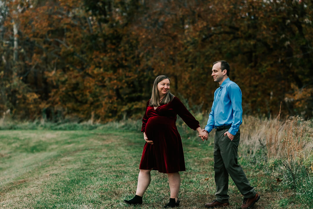 A mom to be in a red velvet dress walks with her husband through a park trail after visiting their postpartum doula dc