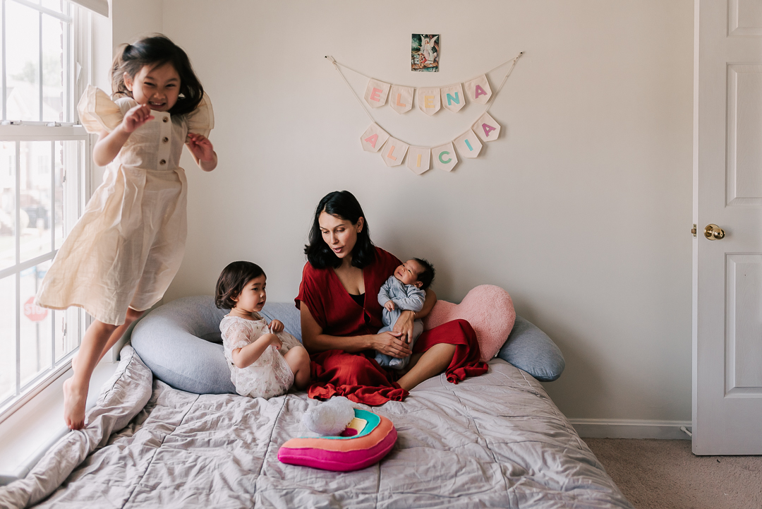 A young girl jumps and plays while mom and her toddler sister sit at the other end of the bed with a newborn baby in mom's lap