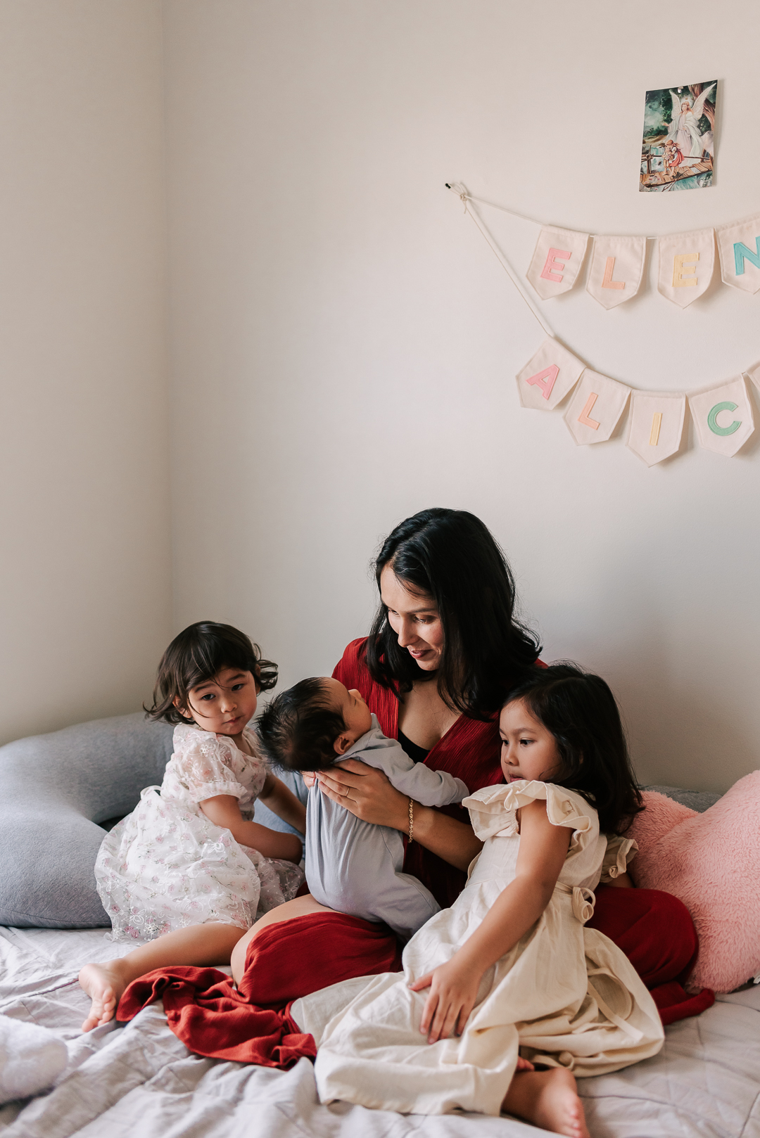 A mother in a red dress sits on a bed with her two toddler daughters playing with a newborn baby thanks to thrive breastfeeding