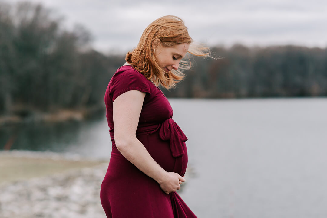 A mother to be in a red maternity dress stands on a windy lake smiling down to her bump after meeting with arlington midwives