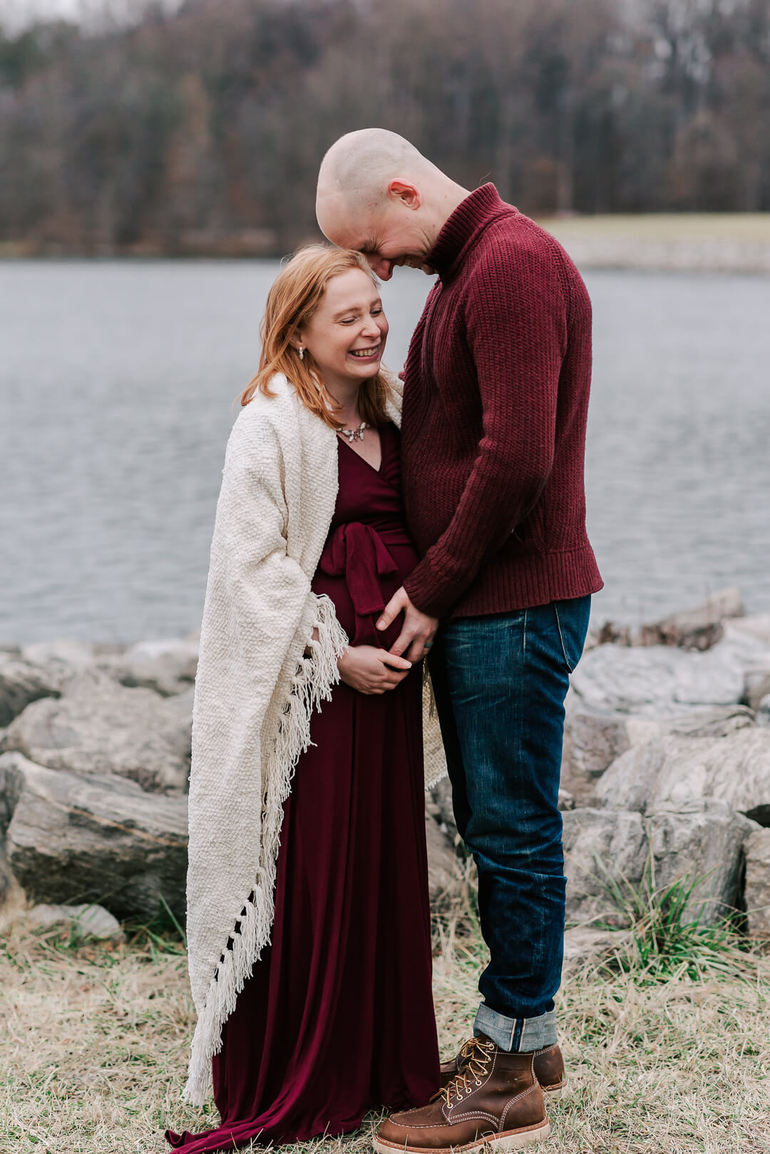 A mother to be laughs with her husband in a red maternity gown as they stand by a lake after meeting arlington midwives