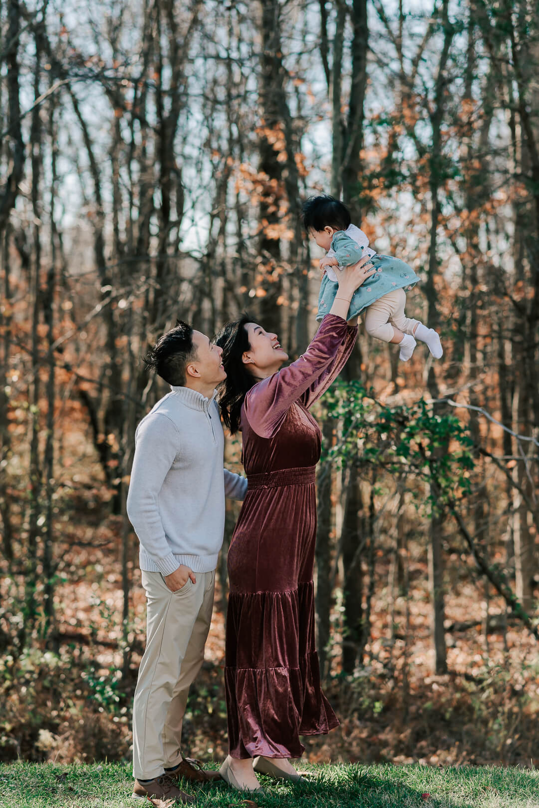A mother in a velvet dress lifts her toddler way over her head with dad looking on in a park after some baby music classes northern virginia