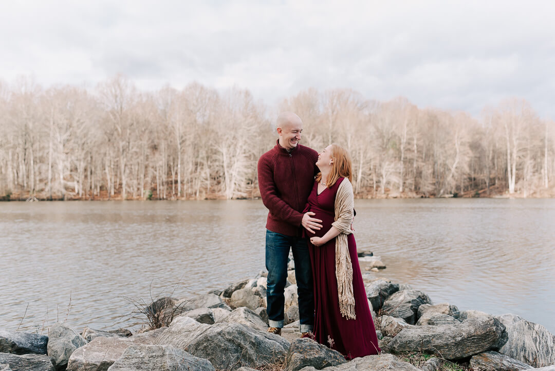 Expecting parents stand on a rock jetty in a lake holding the bump in matching maroon outfits smiling at each other