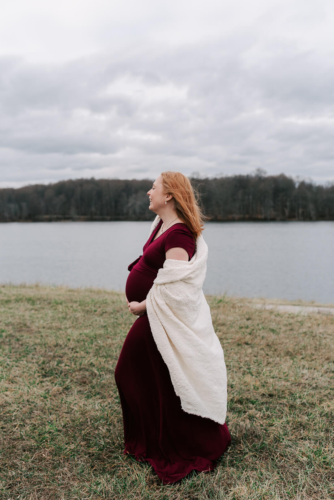 A mother to be walks along a lake in a maroon maternity gown and white blanket after meeting elm and serenity