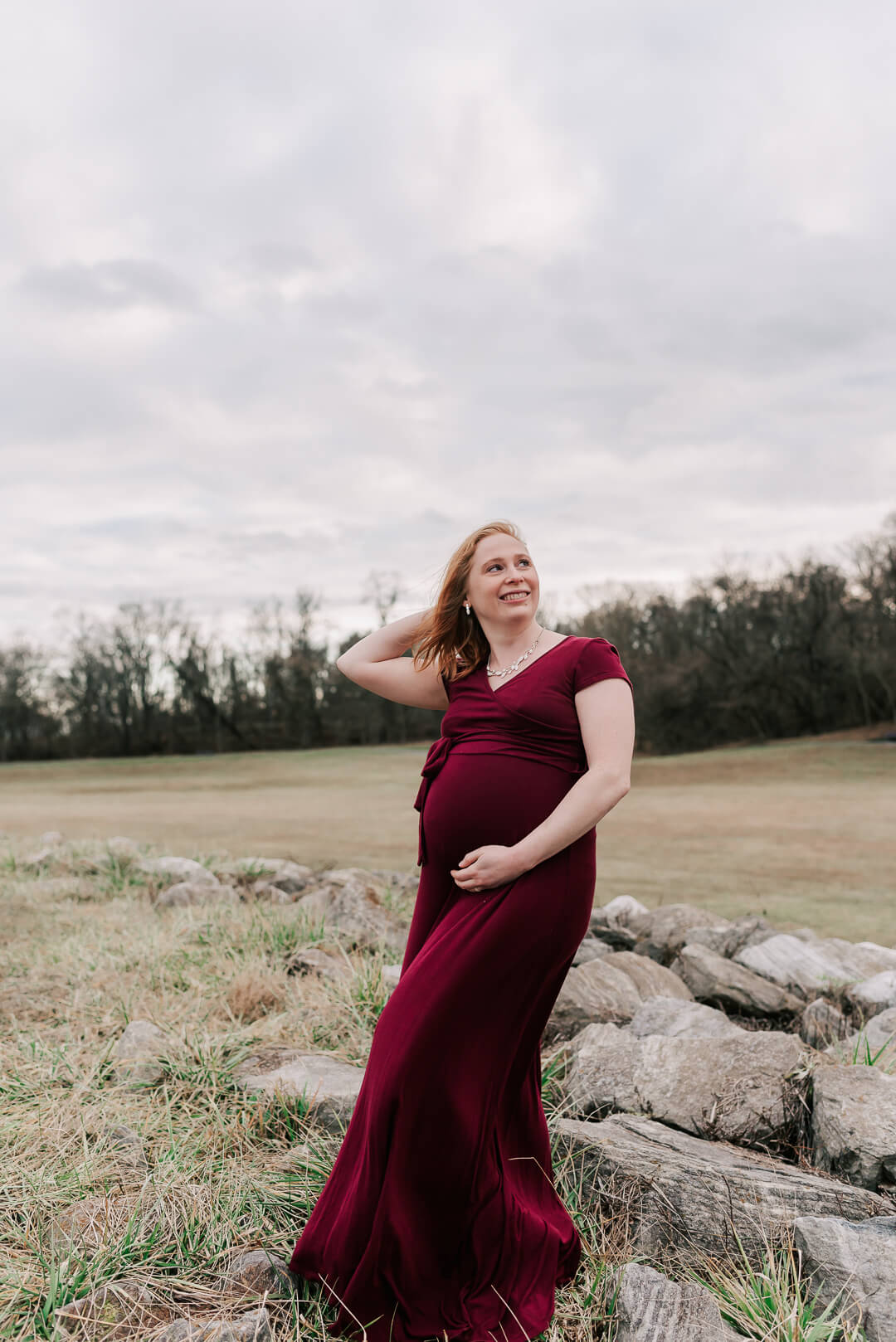 A woman in a maroon maternity dress stands in a field smiling over her shoulder and holding her bump after visiting elm and serenity